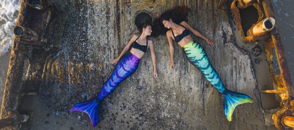 Two girls laying on a ship deck wearing the Hip Flip swimmable mermaid tail. One shows the purple side and the other shows the green and blue side.