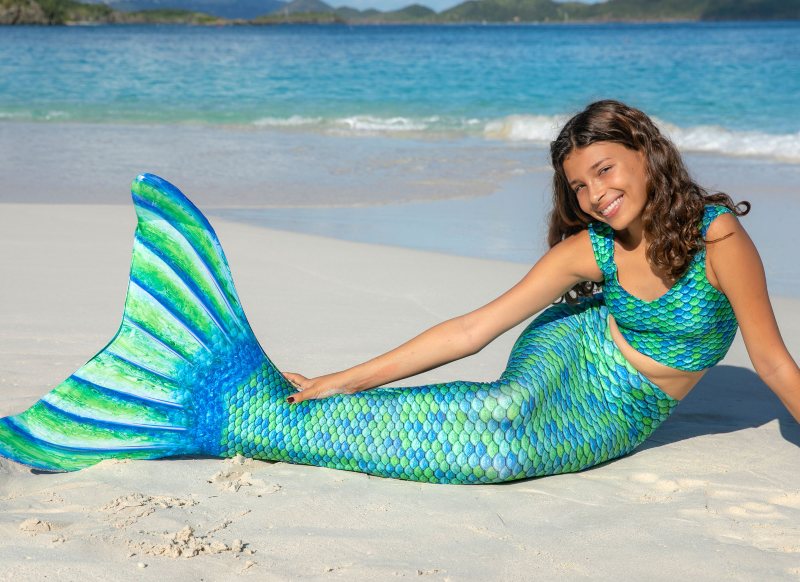 A girl in a blue and green mermaid tail and a matching scaled tankini sitting in the sand.