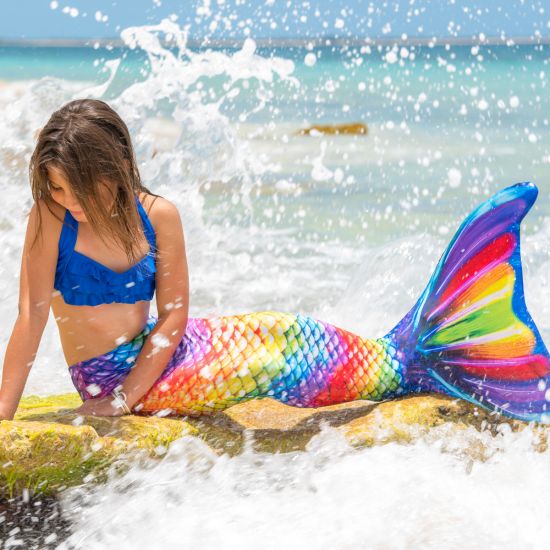 girl on a rock in the Rainbow Reef Mermaidens tail with waves crashing around her