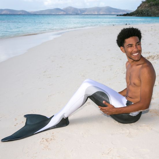 a man in an orca mermaid tail smiles while sitting on the beach