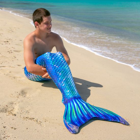 a boy looks out at the ocean while wearing a blue and green mermaid tail and sitting on the beach