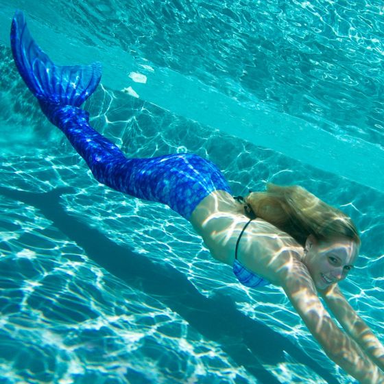 A girl smiling underwater wearing a royal blue swimmable mermaid tail.