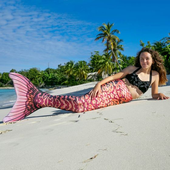 A girl lays on the beach in a coral and orange mermaid tail.