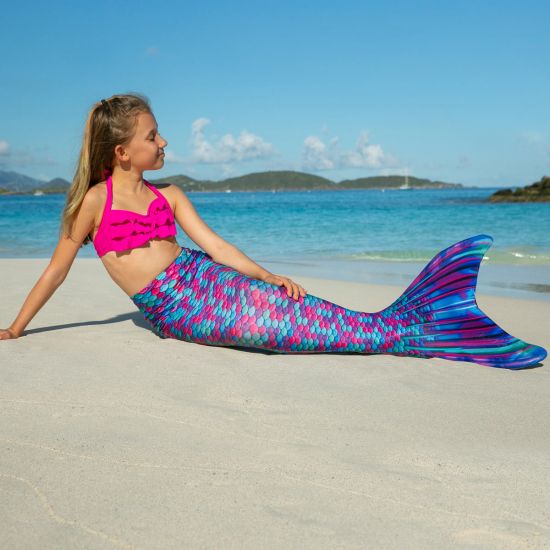 A young girl sits in the sand in a mermaid tail with berry-colored scales.