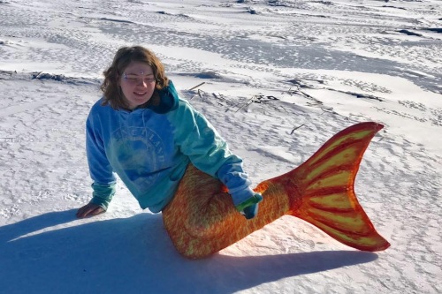 Mermaid Spirit poses in the snow while wearing an orange and yellow mermaid tail.