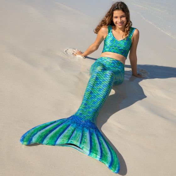 A girl lays in the sand wearing a blue and green-scaled mermaid tail and crop top.