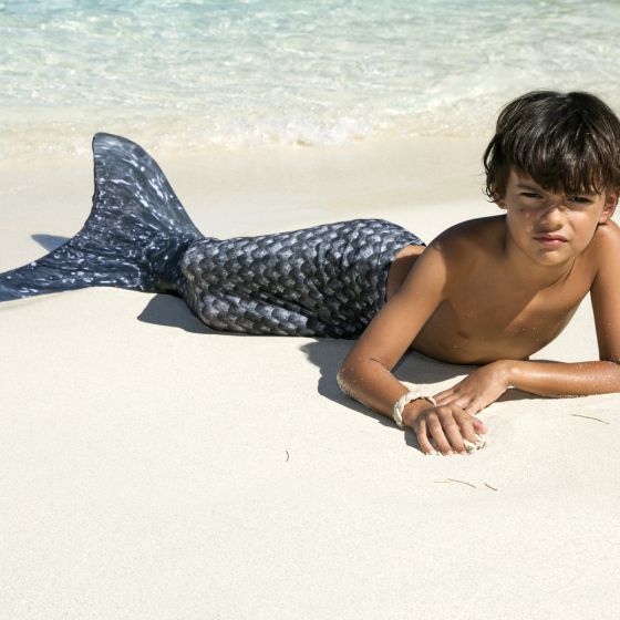 A boy lays in the sand wearing the Barracuda Black merman tail.