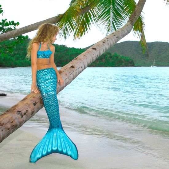 A girl sits on a leaning palm tree in the Tidal Teal mermaid tail.