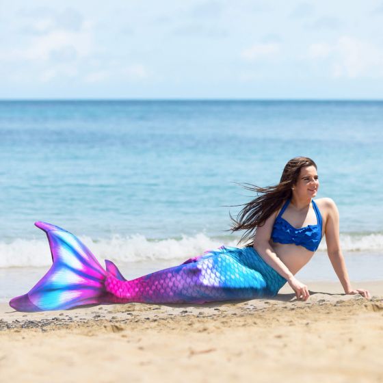 A girl wearing a blue and pink mermaid tail with realistic side, ankle, and dorsal fins lays on her side in the sand.