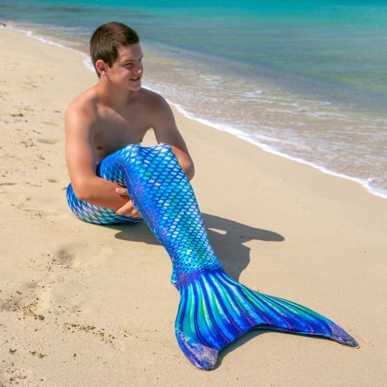 A merman sits on the beach in a green, white, and blue mermaid tail.