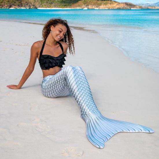 A girl smiles and sits in the sand in a silver swimmable mermaid tail.