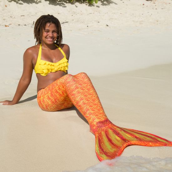 A girl sits in the sand while wearing a yellow and orange mermaid tail and yellow bikini top.