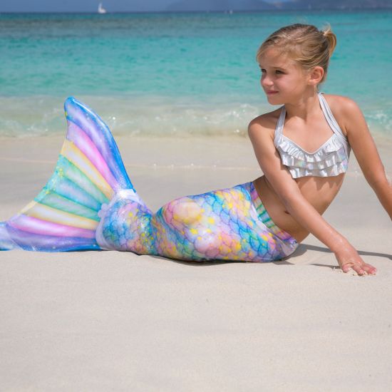 A little girl poses in a pastel mermaid tail next to the ocean.