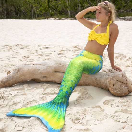 A girl sits on a log while wearing a yellow, green, and blue mermaid tail, a yellow bikini top, and sunglasses.