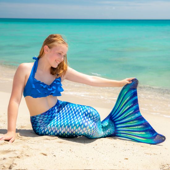 a girls sits in the sand while wearing a blue, green, and white mermaid tail