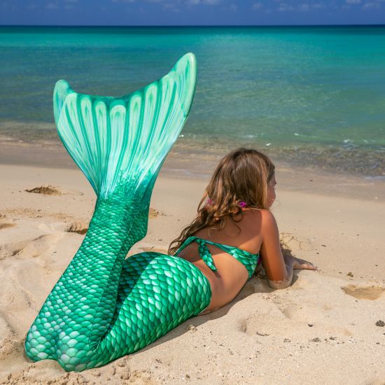 a young mermaid lays on the beach in a green mermaid tail