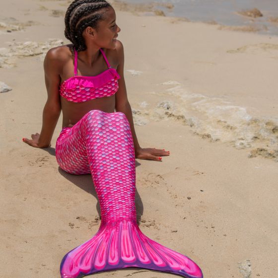 a young mermaid sitting on the beach in a pink mermaid tail