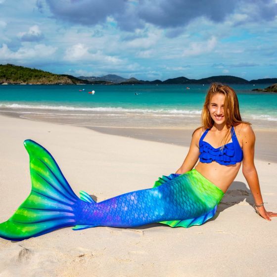 a girl sitting on the beach in a bright green, blue, and purple mermaid tail