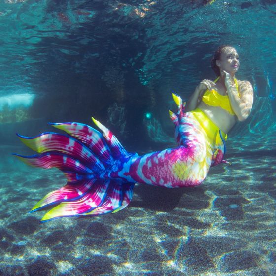 a woman posing underwater in a professional mermaid tail with yellow, pink, blue, and white scales