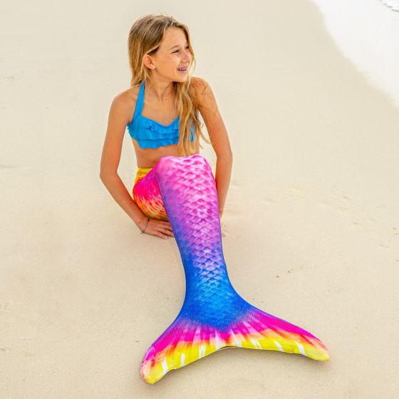 a young girl sitting in the sand in a bright yellow, pink, and blue mermaid tail