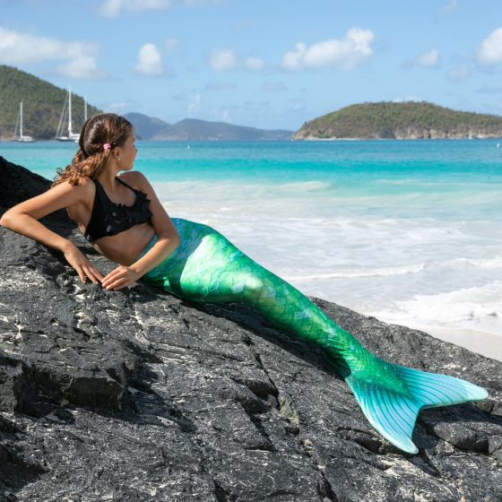 a girl lays on a rocky surface in a green mermaid tail with the ocean in the background